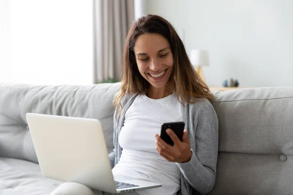 Šťastné tisícileté dívka sedět na gauči multitask pracuje na gadgets — Stock fotografie