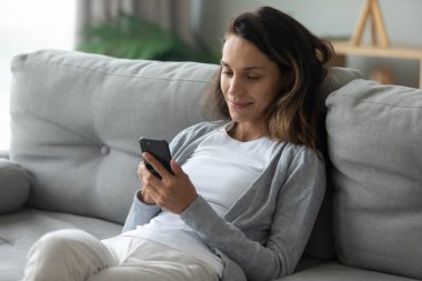 Genç kadın, modern akıllı telefon kullanarak kanepeye oturur.