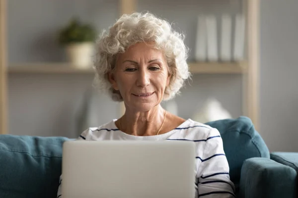 Modern middle-aged 60s woman using laptop relaxing at home