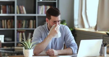 Ill male worker sitting at workplace blowing nose in tissue