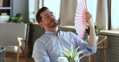 Exhausted young man waving fan suffering from summer heat indoors