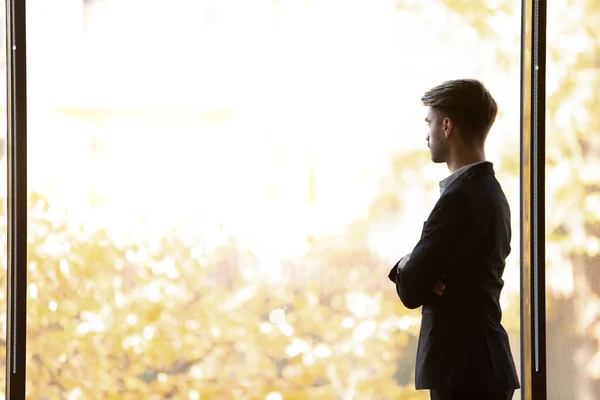 Concentrated thoughtful young businessman looking away at panoramic office window. — Stock Photo, Image