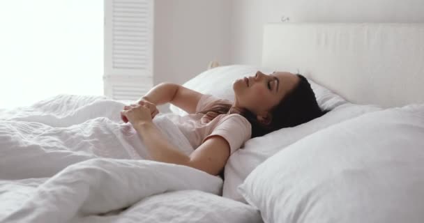 Веселая внимательная молодая женщина просыпается в удобной постели в одиночестве — стоковое видео