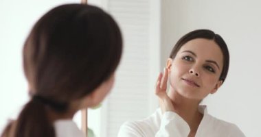 Confident beautiful young woman putting facial cream looking in mirror