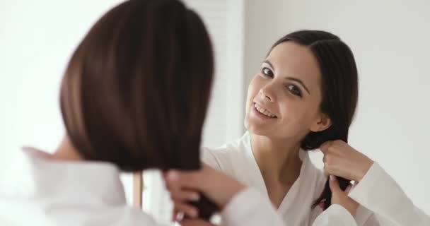 Улыбающаяся молодая красивая женщина касается здоровых волос, глядя в зеркало — стоковое видео