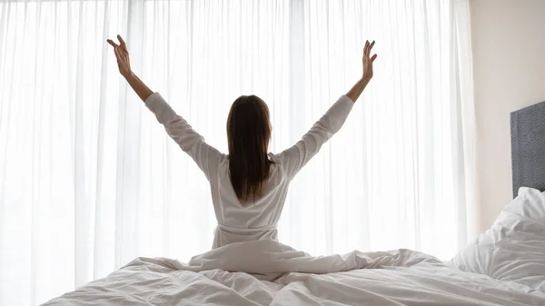 Jongedame in nachtjapon zittend op bed, rijzende armen. — Stockfoto