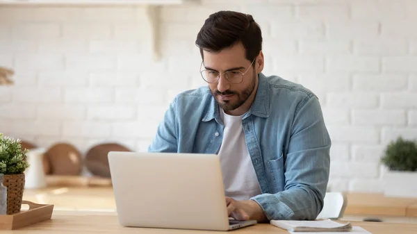 Homem sério usando óculos trabalhando no laptop on-line na cozinha — Fotografia de Stock