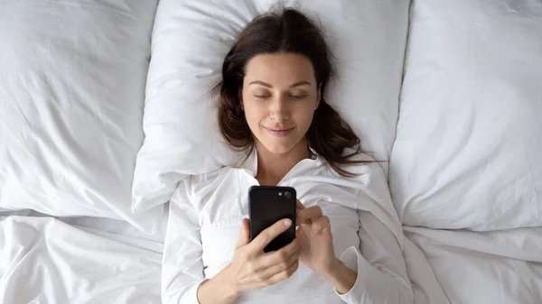 Boldog nő pihenés az ágyban, mobilalkalmazások használata. — Stock Fotó