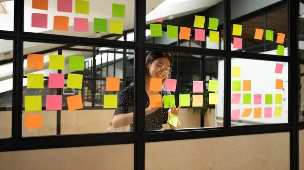 Emocionada empresaria asiática con gafas escribiendo notas en pegatinas —  Fotos de Stock