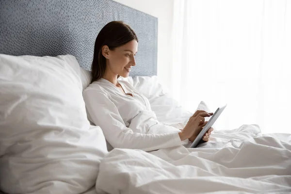 Smiling woman lying in bed under duvet, using digital tablet.
