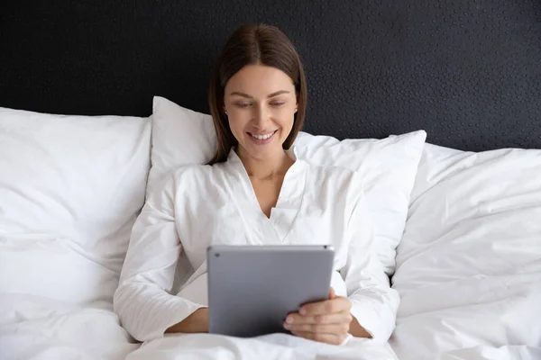 Mujer joven sonriente acostada en una cama cómoda, usando una tableta digital . — Foto de Stock