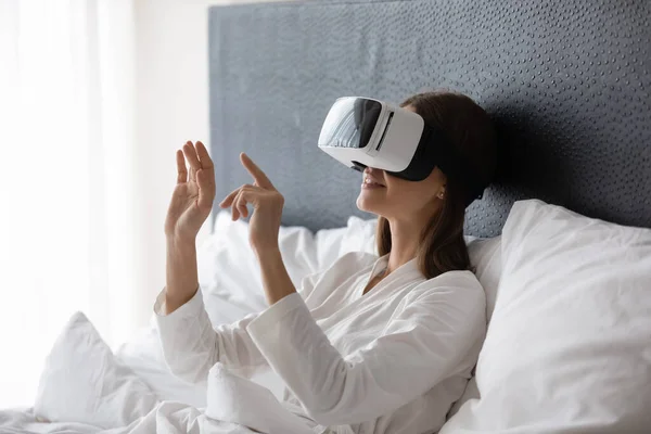 Happy young brunette woman resting in bed, wearing VR googles.