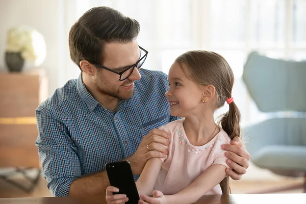 Jovem sorrindo pai olhando para a filha pequena feliz, usando smartphone . — Fotografia de Stock