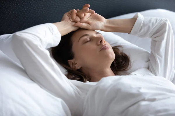 Wanita muda berambut cokelat yang stres terbaring di tempat tidur dengan tangan di atas kepala — Stok Foto