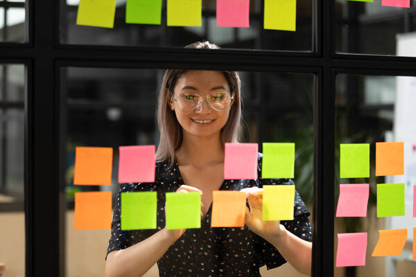 Азиатская предпринимательница пишет заметки на липких бумажках на стеклянной стене
