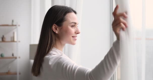 Joven mujer sonriente abriendo la cortina mirando por la ventana — Vídeo de stock