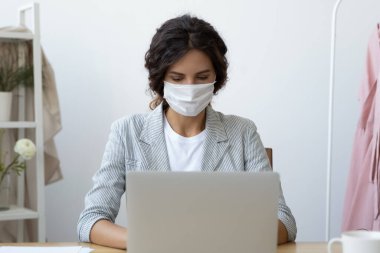 Maskeli bir iş kadını çalışma masasında oturan dizüstü bilgisayarla çalışıyor.