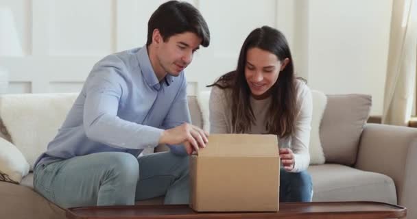 Emocionados clientes jóvenes de la pareja que abren la caja de cartón sentado en el sofá — Vídeo de stock