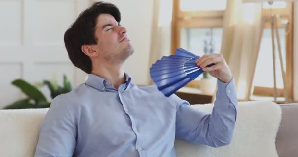 Tired annoyed man waving fan suffering from swelter in apartment — Stock Video