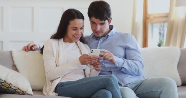 Boldog Y-generációs pár okostelefonnal beszélget a kanapén ülve — Stock videók