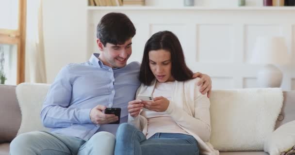 Jóvenes usuarios de la pareja hablando con dos teléfonos relajantes en el sofá — Vídeos de Stock