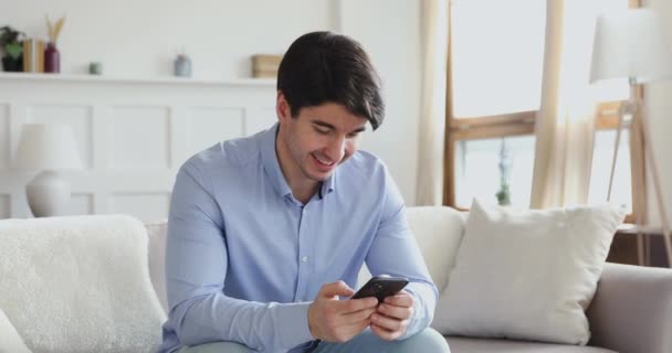 Sorrindo homem milenar usando telefone inteligente sentado no sofá — Vídeo de Stock