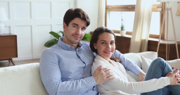 Усміхнена молода пара обіймається сидячи на дивані, дивлячись на камеру — стокове відео
