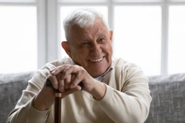 Positive old grandfather holding walking stick resting on couch clipart