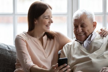 Yetişkin torun ve yaşlı büyükbaba akıllı telefon kullanarak kanepede otururlar.