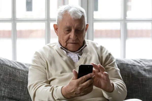 Grey-haired 80s old man sitting on couch with smartphone