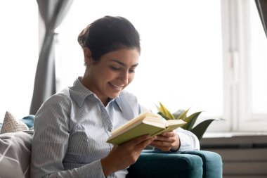 Gülümseyen Hintli kadın kitap okuyor, rahat koltukta oturuyor.