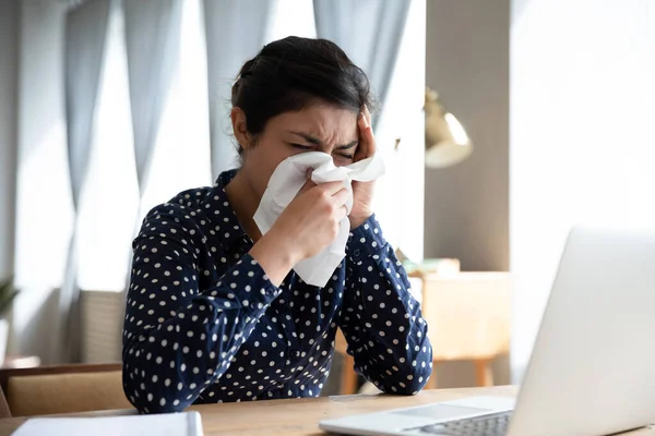 Unhealthy Indian woman blowing nose, sitting at workplace