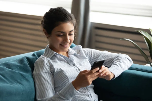 Mujer india sonriente usando un teléfono inteligente, sentada en un sofá acogedor —  Fotos de Stock
