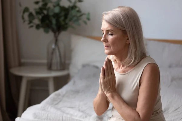 Middle aged woman sitting on bed praying with hope. — Stock Photo, Image