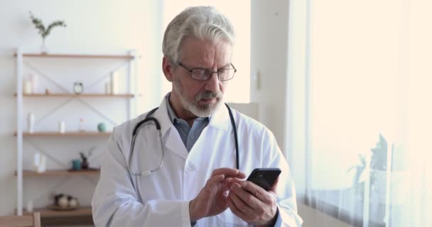 Sorrindo médico sênior segurando smartphone mensagem de texto móvel no escritório — Vídeo de Stock