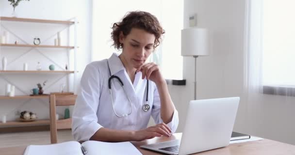 Dizüstü bilgisayar kullanarak beyaz önlük giyen kadın doktor notlar yazıyor. — Stok video