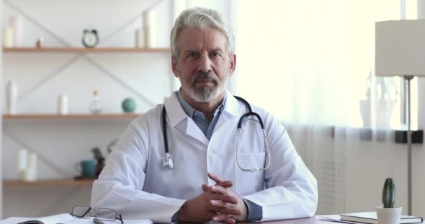 Confiável sorridente profissional mais velho médico sentado no local de trabalho, retrato — Vídeo de Stock