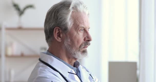 Poważny stary lekarz patrząc przez okno myśląc o wyzwaniach — Wideo stockowe