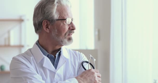 Задумчивый старший врач держит стетоскоп, глядя в окно — стоковое видео