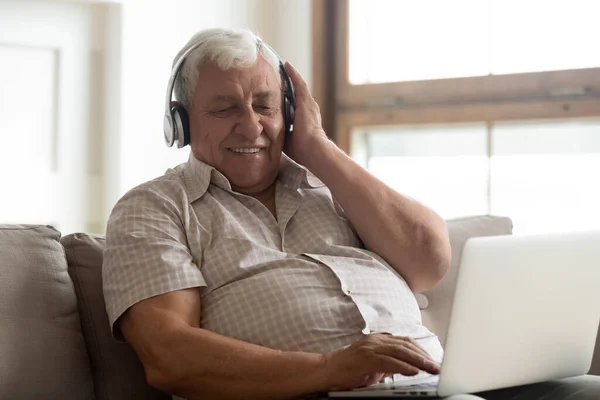 Sonriendo hombre mayor usando auriculares inalámbricos disfrutando de la música, utilizando el ordenador portátil — Foto de Stock