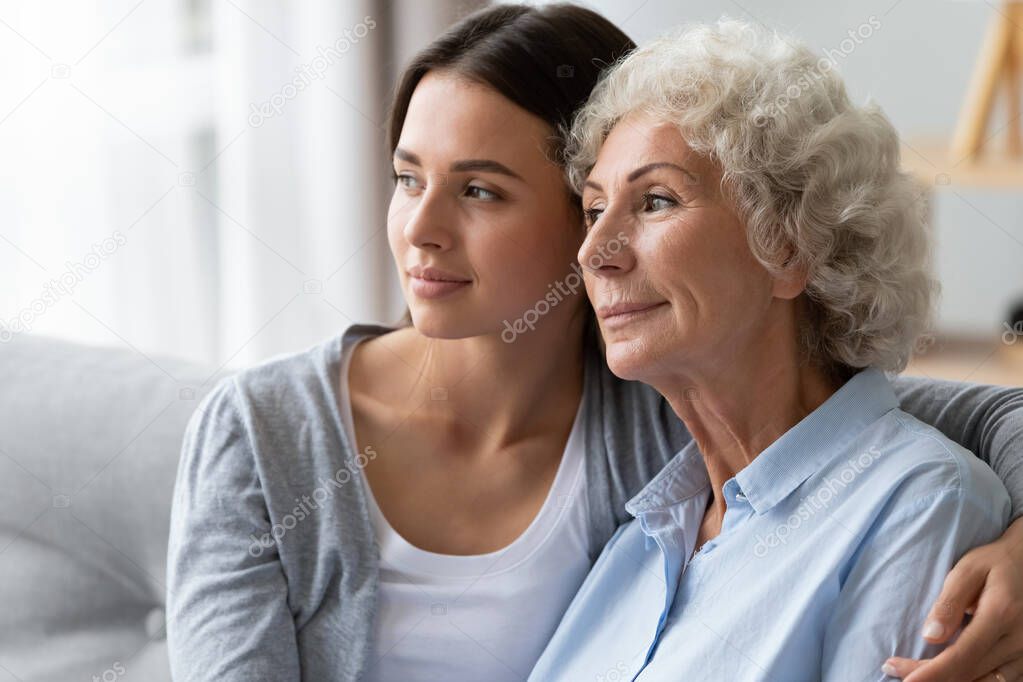 Mature mother and adult daughter cuddle at home thinking