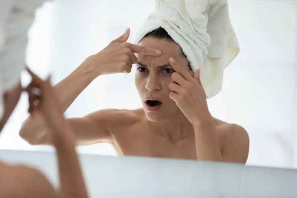 Upset woman looks in mirror squeezes pimple on forehead — Stock Photo, Image