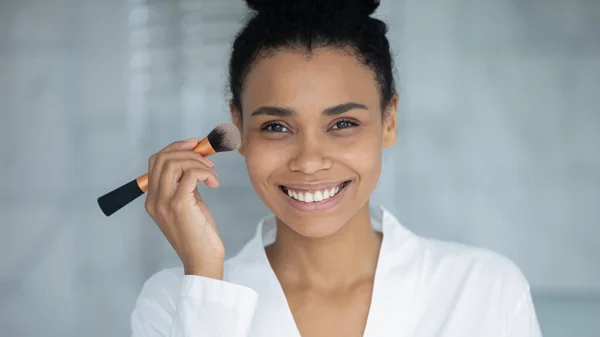 African woman holding brush applying foundation cream on face