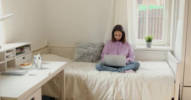 Εστιασμένη έφηβη κοπέλα που φοράει ακουστικά και διαβάζει online στο σπίτι — Αρχείο Βίντεο