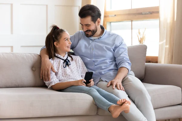 Smiling daddy cuddling pretty cute daughter, using mobile phone. — Stock Photo, Image