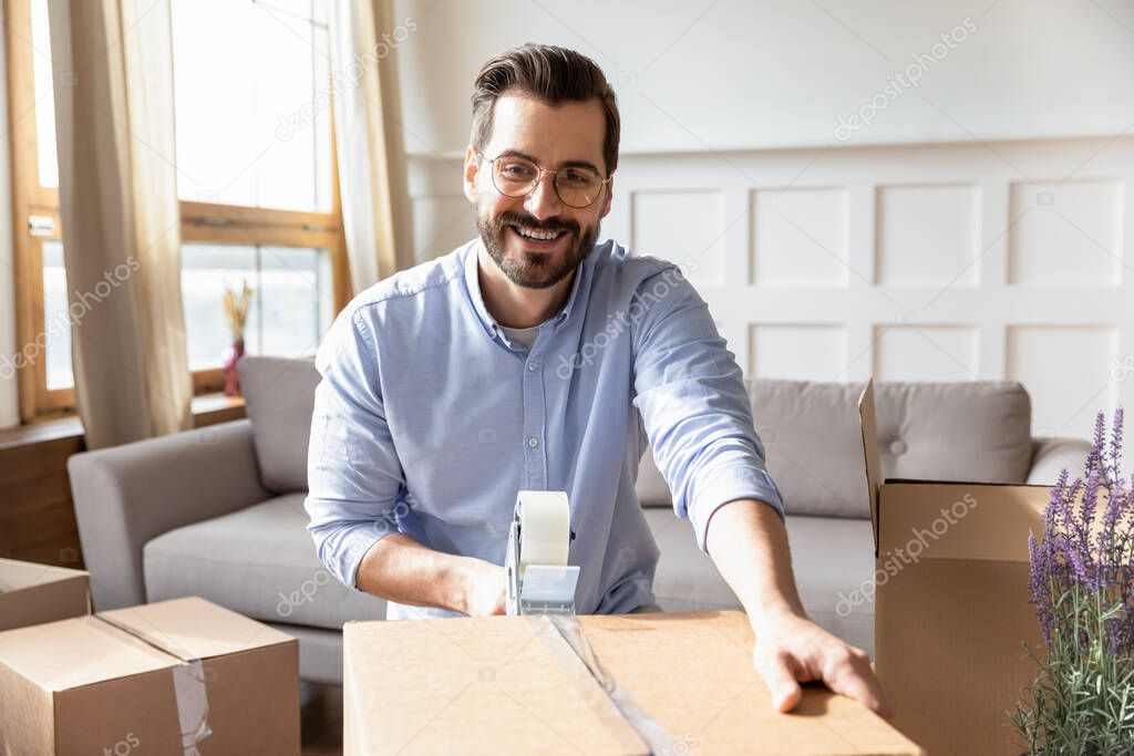 Smiling young bearded man packing personal belongings in cardboard boxes.