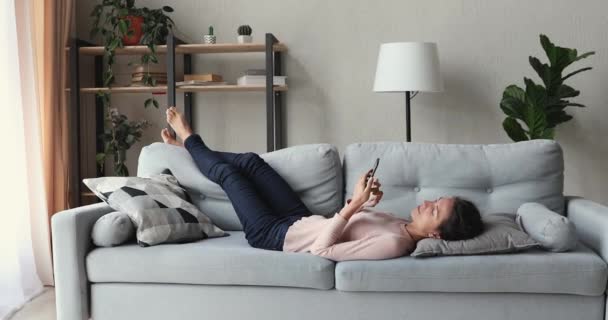 Расслабленная молодая женщина лежит на диване с помощью смартфона — стоковое видео