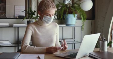 İş kadını sağlık maskesi takıyor. Laptop klavyesini dezenfektanla temizliyor.