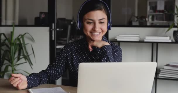Chica india sonriente con auriculares mirando a la computadora portátil haciendo notas — Vídeos de Stock