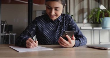 Gülümseyen Hintli genç bayan öğrenci elinde akıllı telefon, notlar alıyor.